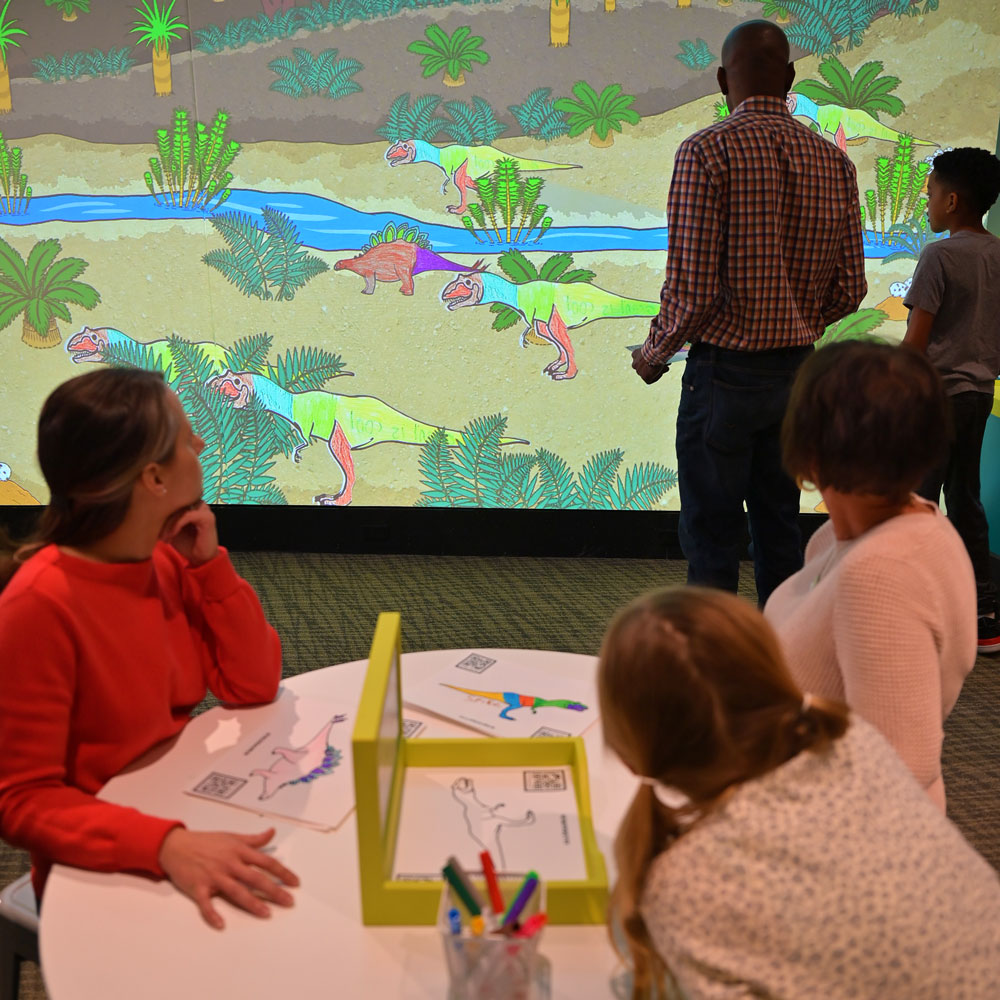 People are sitting at a table with art supplies in the foreground. A child and grown-up are standing and looking for their design on the projected screen in the background.